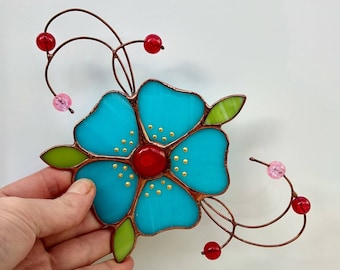 May include: A turquoise blue stained glass flower with a red center and gold accents. The flower is attached to a copper wire stem with red and pink glass beads.