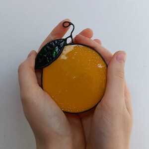 May include: A stained glass orange with a green leaf and black outline. The orange is a vibrant yellow with white speckles. The leaf is a dark green. The orange is held in cupped hands.