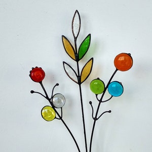 Puede incluir: Una escultura floral artesanal de metal y vidrio. La escultura presenta un tallo central con globos de vidrio de colores en rojo, naranja, verde, amarillo y azul. Las hojas son amarillas, verdes y blancas. El metal es oscuro.