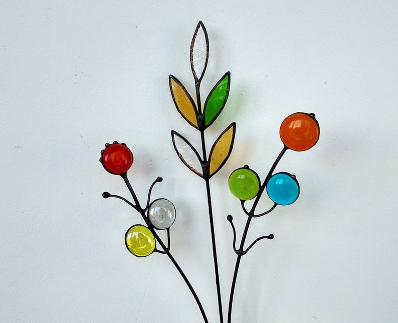 Puede incluir: Una escultura floral artesanal de metal y vidrio. La escultura presenta un tallo central con globos de vidrio de colores en rojo, naranja, verde, amarillo y azul. Las hojas son amarillas, verdes y blancas. El metal es oscuro.