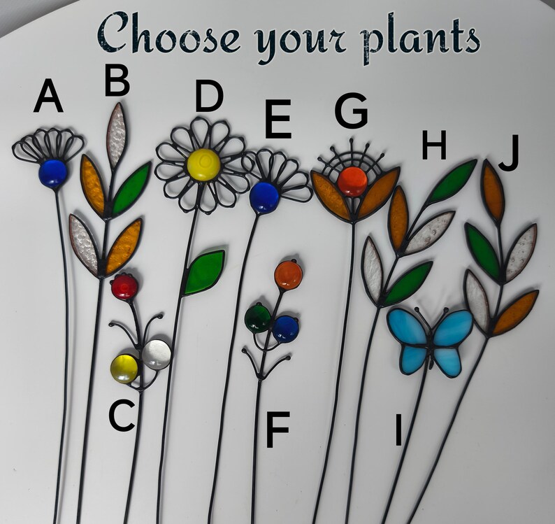 Puede incluir: Una selecci&oacute;n de estacas decorativas para jard&iacute;n con acentos de vidrio de colores. Los dise&ntilde;os incluyen flores, hojas y una mariposa. El texto "Choose your plants" est&aacute; en la parte superior. Cada estaca tiene un tallo de metal negro.