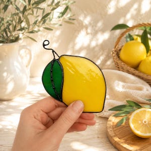May include: A stained glass lemon with a green leaf and black outline. The lemon is yellow and the leaf is green. The glass art is held in a hand, with lemons and a basket in the background.