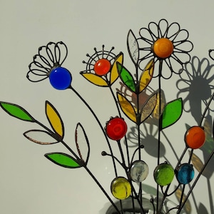 Puede incluir: Una escultura floral de metal con detalles de vidrio de colores. Las flores presentan cuentas y p&eacute;talos de vidrio amarillo, naranja, rojo, azul y verde. Los tallos son oscuros y las hojas est&aacute;n delineadas en negro.