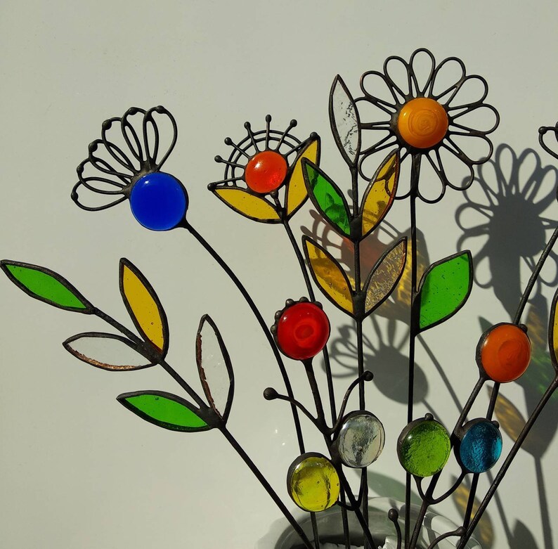 Puede incluir: Una escultura floral de metal con detalles de vidrio de colores. Las flores presentan cuentas y p&eacute;talos de vidrio amarillo, naranja, rojo, azul y verde. Los tallos son oscuros y las hojas est&aacute;n delineadas en negro.