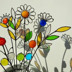Puede incluir: Un arreglo decorativo de flores de metal y vidrio. Las flores presentan diversas cuentas y hojas de vidrio de colores, con una flor en forma de margarita en el centro. Los tallos son de metal oscuro y las flores proyectan sombras sobre el fondo blanco.