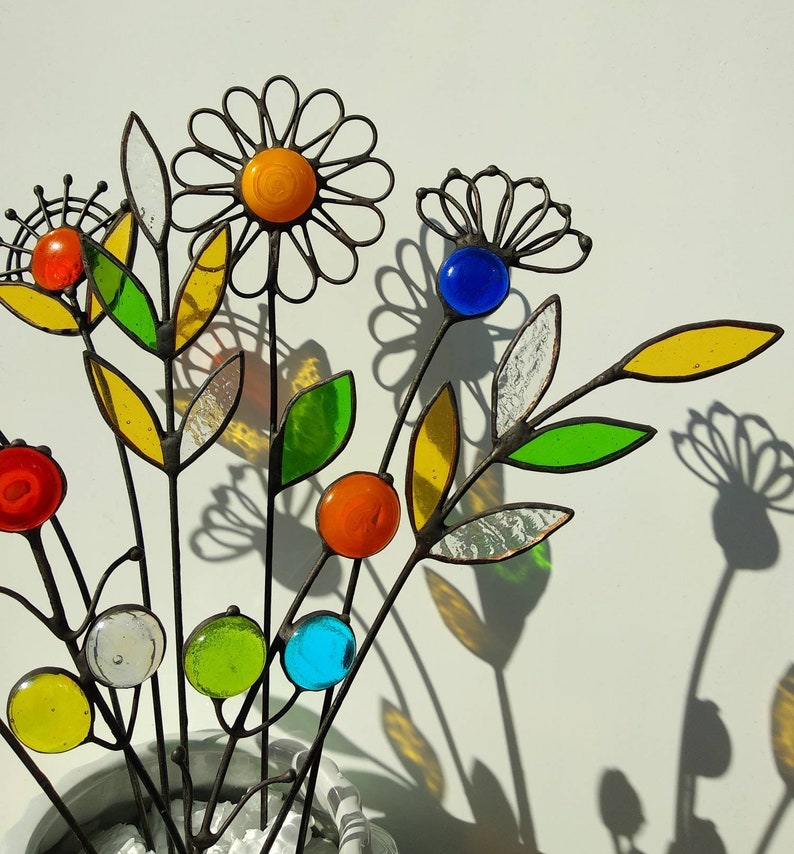 Puede incluir: Un arreglo decorativo de flores de metal y vidrio. Las flores presentan diversas cuentas y hojas de vidrio de colores, con una flor en forma de margarita en el centro. Los tallos son de metal oscuro y las flores proyectan sombras sobre el fondo blanco.