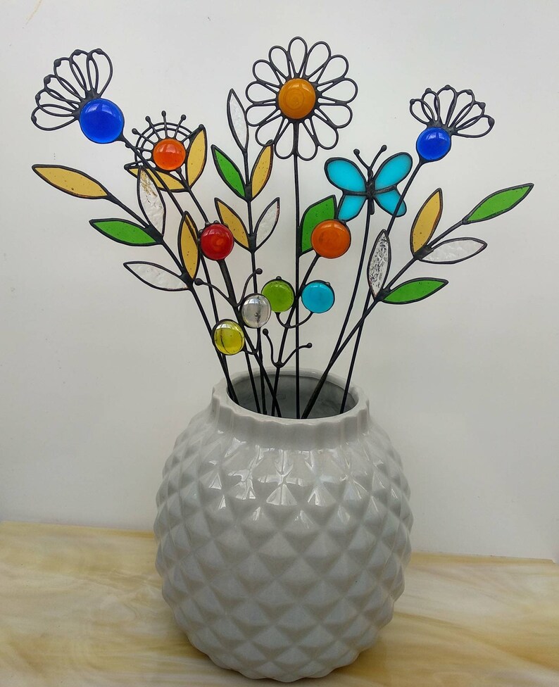 Puede incluir: Un jarr&oacute;n de cer&aacute;mica gris con un patr&oacute;n de diamantes contiene un ramo de flores decorativas de metal. Las flores presentan detalles de vidrio de colores en azul, naranja, amarillo y verde, con un dise&ntilde;o de mariposa y margarita.