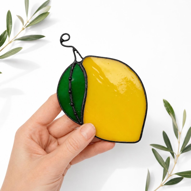 May include: A stained glass lemon with a green leaf and black wire detailing. The lemon is a bright yellow color, and the leaf is a vibrant green. The piece is held in a hand, with olive branches in the background.