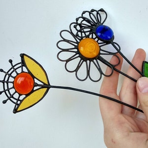 Puede incluir: Estacas decorativas de jard&iacute;n con forma de flor de metal con detalles de vidrio de colores. Una flor tiene un centro y p&eacute;talos amarillos, con un orbe de vidrio naranja. Otra tiene un orbe de vidrio azul y un centro amarillo. Los tallos son negros.