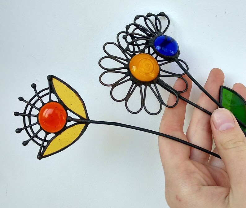 Puede incluir: Estacas decorativas de jard&iacute;n con forma de flor de metal con detalles de vidrio de colores. Una flor tiene un centro y p&eacute;talos amarillos, con un orbe de vidrio naranja. Otra tiene un orbe de vidrio azul y un centro amarillo. Los tallos son negros.