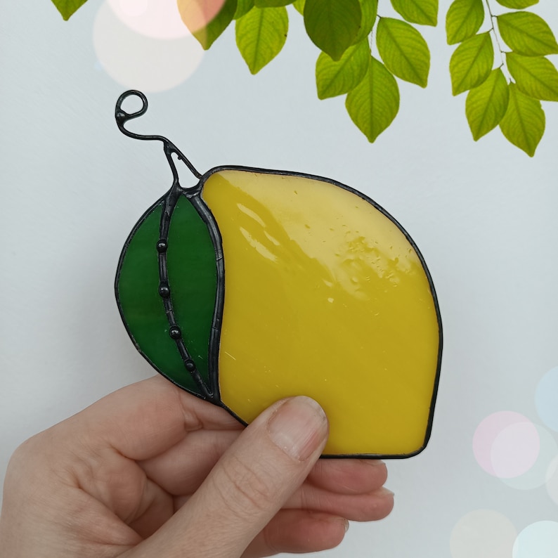 May include: A stained glass lemon with a green leaf and black outline. The lemon is yellow and has a curved shape. The leaf is green with black dots. The lemon is held in a hand.
