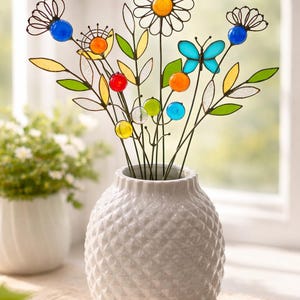 Puede incluir: Un jarr&oacute;n blanco texturizado contiene un colorido arreglo de flores y hojas de metal decorativas. El arreglo incluye una margarita, una mariposa y varias cuentas de vidrio de colores. Las flores son de metal y vidrio.