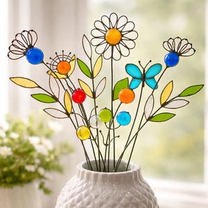 Puede incluir: Un ramo de flores de cristal de colores y una mariposa en un jarr&oacute;n blanco. Las flores tienen detalles amarillos, naranjas, rojos, azules y verdes. Las hojas son verdes y amarillas. El jarr&oacute;n tiene una superficie texturizada.