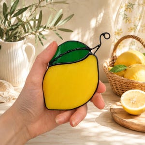 May include: A stained glass lemon with a green leaf and black outline. The lemon is yellow with a green leaf and a black wire stem. The lemon is held in a hand, with a basket of lemons in the background.