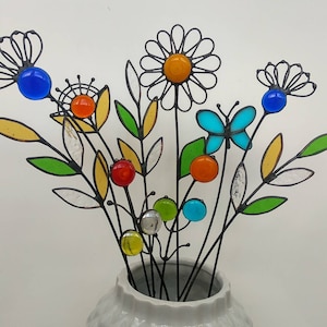 Puede incluir: Un ramo de flores y hojas de metal coloridas hechas a mano en un jarr&oacute;n blanco. Las flores presentan vibrantes detalles de vidrio en azul, naranja, amarillo y verde. Tambi&eacute;n se incluye una mariposa.