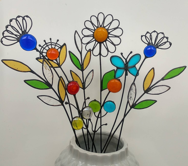 Puede incluir: Un ramo de flores y hojas de metal coloridas hechas a mano en un jarr&oacute;n blanco. Las flores presentan vibrantes detalles de vidrio en azul, naranja, amarillo y verde. Tambi&eacute;n se incluye una mariposa.