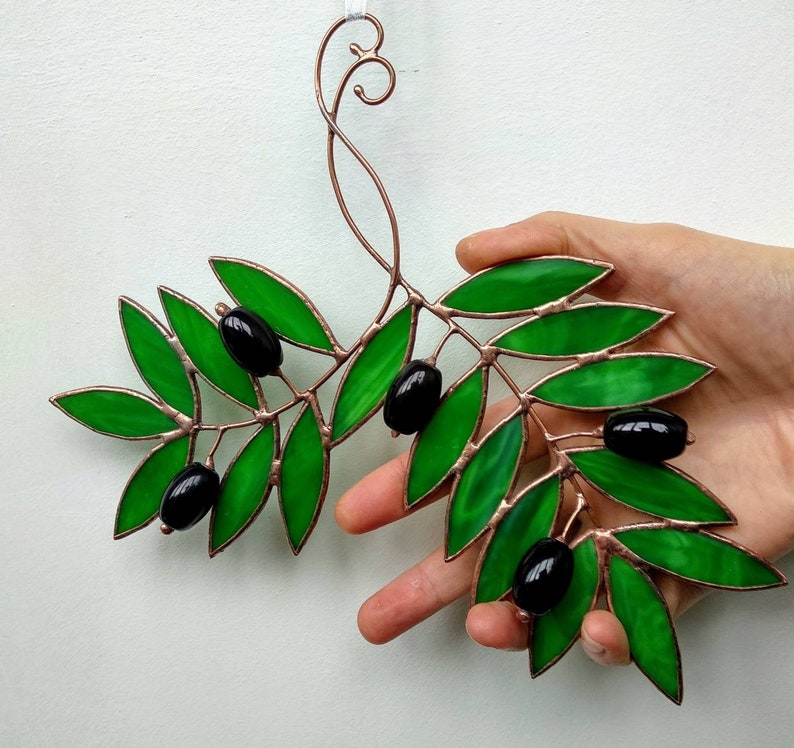 K&ouml;nnte beinhalten: Ein Olivenzweig aus Glasmalerei mit gr&uuml;nen Bl&auml;ttern und schwarzen Oliven. Die Bl&auml;tter sind ein leuchtendes Gr&uuml;n, und die Oliven sind gl&auml;nzend schwarz. Der kupferfarbene Rahmen verleiht einen Hauch von Eleganz. Ein dekoratives St&uuml;ck f&uuml;r die Inneneinrichtung.