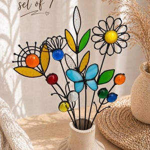 Puede incluir: Un conjunto de siete flores y mariposas decorativas de vidrieras en un jarr&oacute;n crema. Las flores tienen detalles en amarillo, naranja, azul y verde. El texto "set of 7" est&aacute; escrito en cursiva en la parte superior.