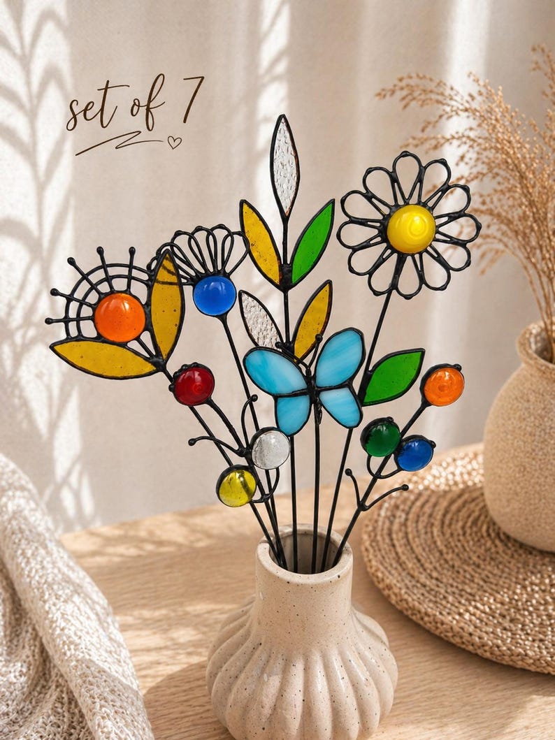 Puede incluir: Un conjunto de siete flores y mariposas decorativas de vidrieras en un jarr&oacute;n crema. Las flores tienen detalles en amarillo, naranja, azul y verde. El texto "set of 7" est&aacute; escrito en cursiva en la parte superior.