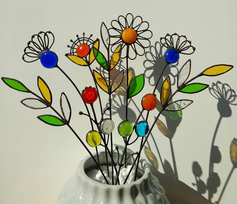 Puede incluir: Un ramo de flores de metal coloridas en un jarr&oacute;n blanco. Las flores presentan p&eacute;talos de vidrio y acentos esf&eacute;ricos en tonos azules, naranjas, amarillos, rojos y verdes. Los tallos son de metal oscuro y las hojas son de vidrieras.