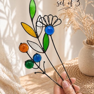 Puede incluir: Juego de tres estacas decorativas florales para jard&iacute;n. Cada estaca presenta p&eacute;talos y orbes de vidrio de colores en tonos verdes, amarillos, naranjas y azules, con tallos de metal negro. El texto "set of 3" est&aacute; escrito en cursiva.