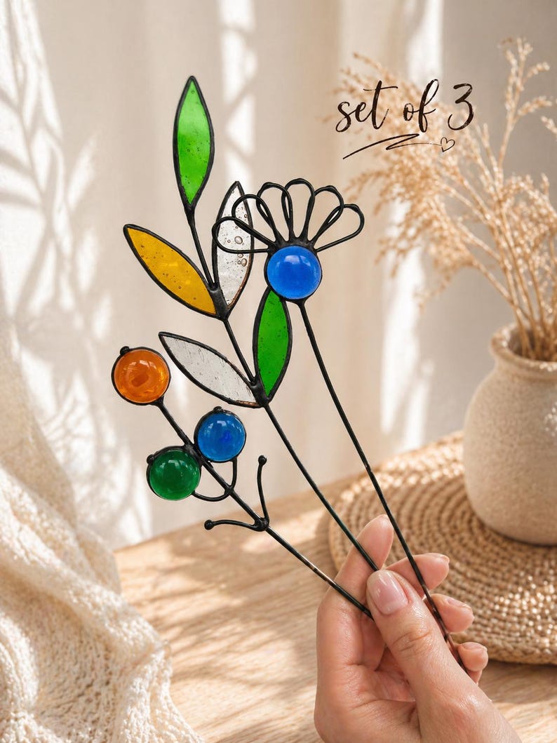Puede incluir: Juego de tres estacas decorativas florales para jard&iacute;n. Cada estaca presenta p&eacute;talos y orbes de vidrio de colores en tonos verdes, amarillos, naranjas y azules, con tallos de metal negro. El texto "set of 3" est&aacute; escrito en cursiva.