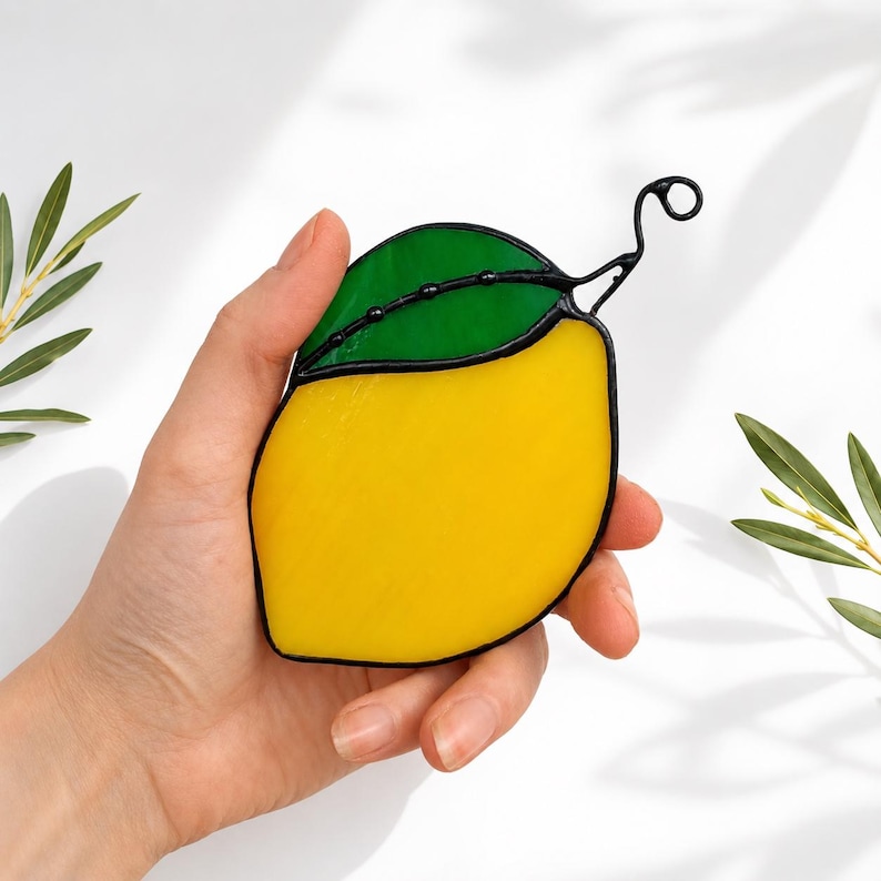May include: A stained glass lemon with a green leaf and black outlines. The lemon is yellow, and the leaf is green. The lemon is held in a hand against a white background. The lemon has a curly stem.