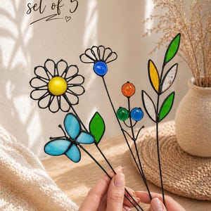 Puede incluir: Un conjunto de cinco estacas de jard&iacute;n de vidrieras de colores. Las estacas presentan una margarita, una mariposa y otros dise&ntilde;os florales con acentos azules, amarillos, naranjas y verdes. El texto "set of 5" est&aacute; escrito en cursiva.