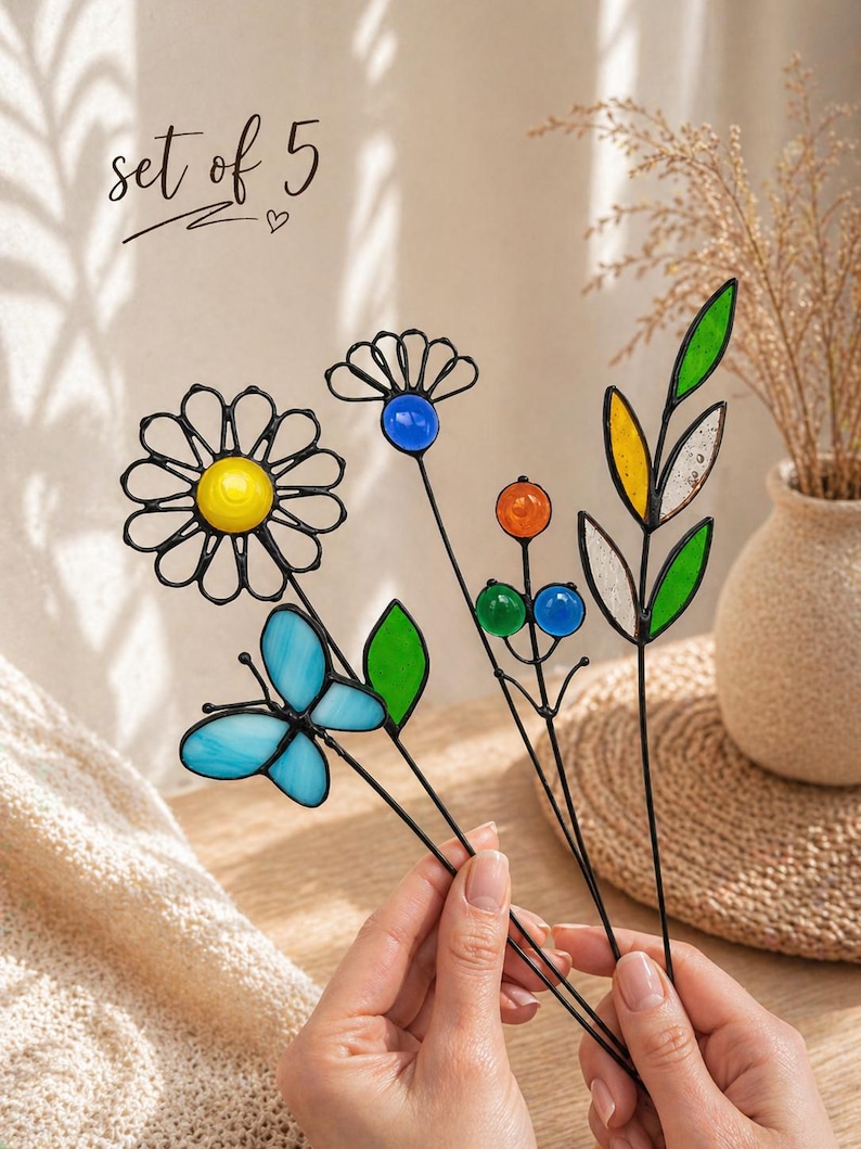 Puede incluir: Un conjunto de cinco estacas de jard&iacute;n de vidrieras de colores. Las estacas presentan una margarita, una mariposa y otros dise&ntilde;os florales con acentos azules, amarillos, naranjas y verdes. El texto "set of 5" est&aacute; escrito en cursiva.