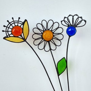 Puede incluir: Tres estacas de jard&iacute;n de metal con dise&ntilde;os de flores de vidrio de colores. Las flores presentan centros de vidrio naranja, amarillo y azul, con contornos de metal negro. Una tiene un p&eacute;talo amarillo, otra una hoja verde.