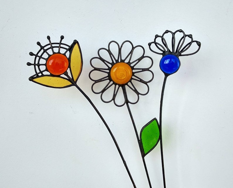 Puede incluir: Tres estacas de jard&iacute;n de metal con dise&ntilde;os de flores de vidrio de colores. Las flores presentan centros de vidrio naranja, amarillo y azul, con contornos de metal negro. Una tiene un p&eacute;talo amarillo, otra una hoja verde.