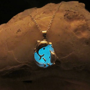 May include: A silver necklace with a pendant depicting dolphins. The pendant has a blue, glowing centre with dolphin silhouettes. The necklace is displayed on a textured, light brown surface.