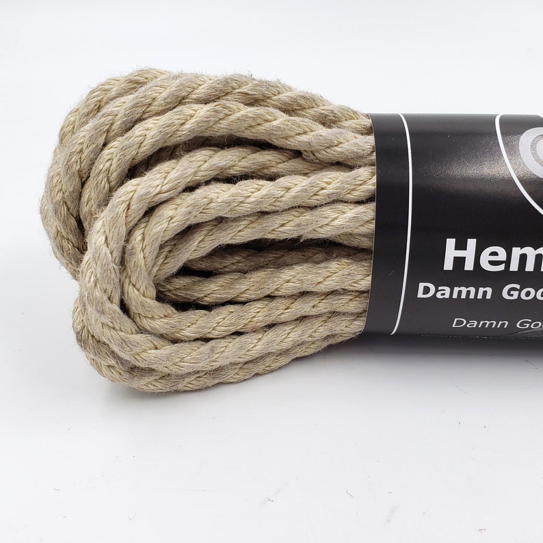 May include: A tightly coiled bundle of natural hemp rope. The rope is a light tan color and has a textured, twisted appearance. A black label with white text reads "Hemp Damn Good".