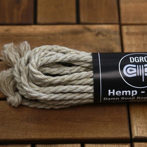 May include: A bundle of natural hemp rope, 6mm thick, with a black label that reads "Hemp - 6mm" and "Damn Good Rope Company." The rope is beige and tightly wound, with frayed ends. The background is a wooden surface.