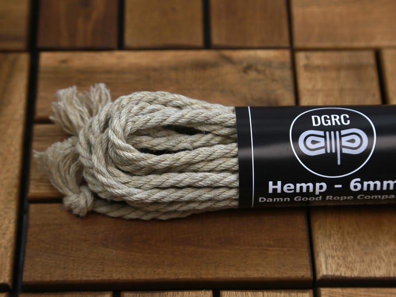 May include: A bundle of natural hemp rope, 6mm thick, with a black label that reads "Hemp - 6mm" and "Damn Good Rope Company." The rope is beige and tightly wound, with frayed ends. The background is a wooden surface.