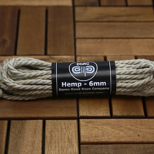 May include: A coil of natural hemp rope, 6mm in diameter, with a black label that reads "Hemp - 6mm" and "Damn Good Rope Company". The rope is beige and tightly wound. The background is a wooden surface.