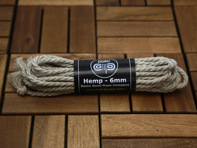 May include: A coil of natural hemp rope, 6mm in diameter, with a black label that reads "Hemp - 6mm" and "Damn Good Rope Company". The rope is beige and tightly wound. The background is a wooden surface.