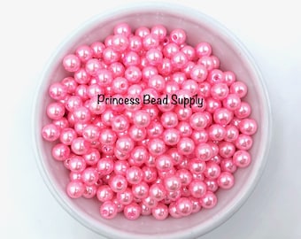May include: A white bowl filled with numerous pink, round beads. The beads have a glossy finish and are labeled "Princess Bead Supply". These beads are suitable for jewelry making and crafting projects.