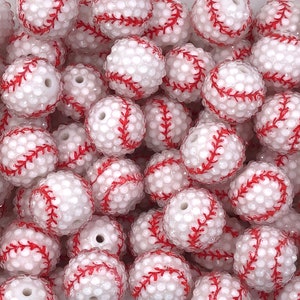 May include: A collection of white baseball-themed beads with red stitching. The beads have a textured surface and are designed to resemble baseballs. These beads are suitable for crafting projects.