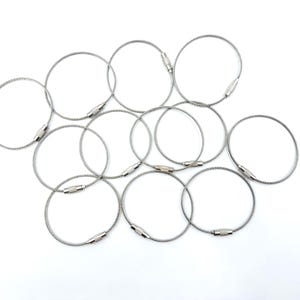 May include: Ten silver cable key rings with a silver metal clasp. The key rings are arranged in a circle on a white background.