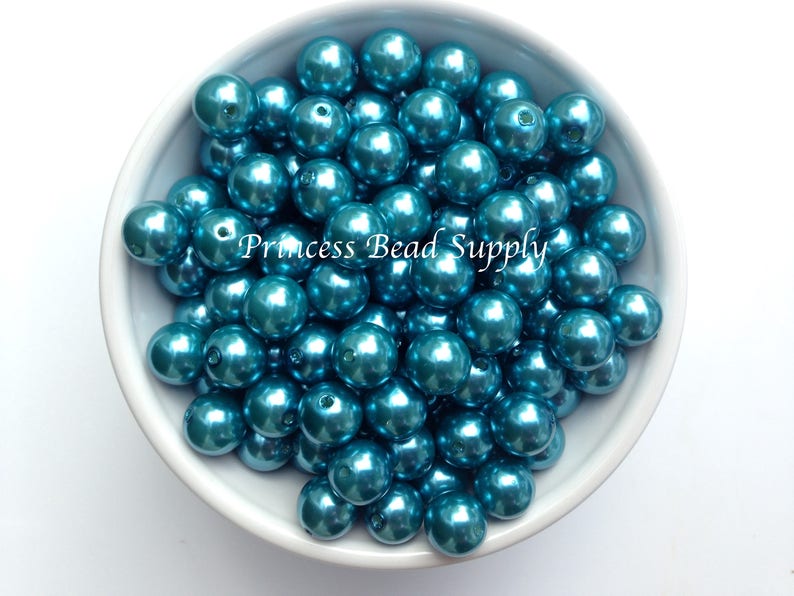 May include: A bowl filled with teal blue pearl-like beads. The beads are smooth and shiny, and they have a metallic finish. The beads are all the same size and shape.