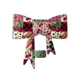 May include: A decorative bow with a patchwork design featuring cherries, stars, and checkered patterns in pink, red, green, and white. The bow has a soft, plush texture and is likely used for hair accessories or gift wrapping.