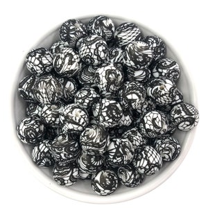 May include: A white bowl filled with black and white patterned beads. The beads have a round shape and are decorated with a black lace-like design. The beads are a craft supply.
