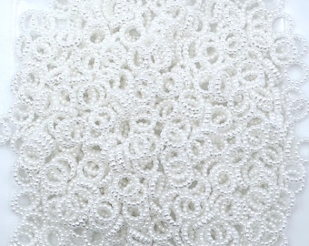 May include: A pile of small, white, beaded rings. Each ring is made of tiny, round, white beads, creating a delicate, textured appearance. The rings are closely packed together, filling the frame.