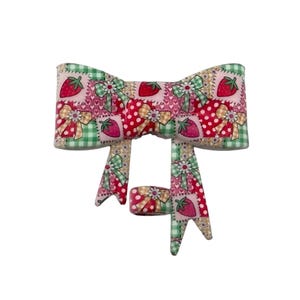 May include: A fabric bow with a patchwork design featuring strawberries, polka dots, and floral accents. The bow has a mix of red, pink, green, and cream colors. The bow is a decorative accessory.