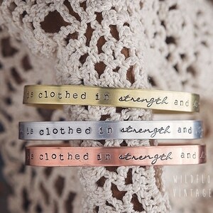 May include: Three stacked metal bracelets with the inscription "is clothed in strength and" on each. The bracelets are gold, silver, and copper.