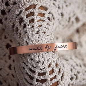 May include: A copper-colored cuff bracelet with the words "walk by faith" engraved in a handwritten style. The bracelet is set against a backdrop of a cream-colored, open-weave fabric. The Wildflower Vintage logo is visible.