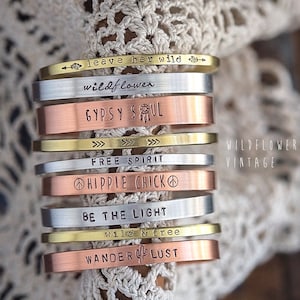May include: A stack of eight silver, gold, and copper bracelets with various inspirational phrases engraved on them, including "leave her wild", "wildflower", "gypsy soul", "free spirit", "hippie chick", "be the light", "wild & free", and "wanderlust".