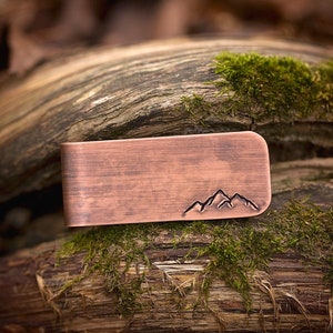 May include: A copper money clip with a mountain range design. The clip has a brushed finish and is rectangular in shape.