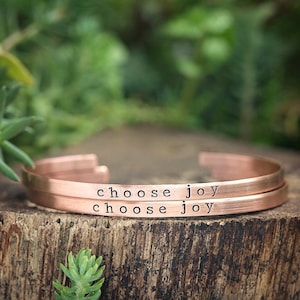 May include: Two copper bracelets with the words "choose joy" engraved on them. The bracelets are stacked on top of each other on a wooden surface.